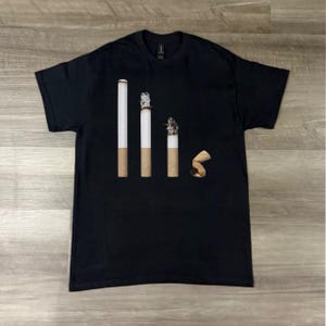 Puede incluir: Camiseta negra con un gráfico que representa un cigarrillo en diferentes etapas de ser fumado. El diseño muestra un cigarrillo lleno, uno parcialmente fumado, uno casi terminado y una colilla desechada.