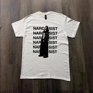 Puede incluir: Una camiseta blanca de manga corta con un estampado gráfico negro de una persona con chaqueta y pantalones. La palabra NARCISSIST se repite seis veces en letras mayúsculas negras detrás de la figura, con las letras S-I-S-T parcialmente ocultas.