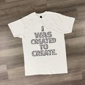 Puede incluir: Camiseta blanca con el texto "I WAS CREATED TO CREATE" en un diseño de estampado de leopardo. La camiseta está hecha de un material suave y tiene un cuello redondo clásico.