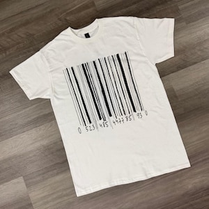 Könnte beinhalten: Weißes T-Shirt mit einem schwarzen Barcode-Design. Der Barcode besteht aus vertikalen Linien und enthält die Zahlen 0 523 485 4477 85 93 0 am unteren Rand. Das T-Shirt hat kurze Ärmel und einen Rundhalsausschnitt.