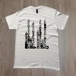 May include: White t-shirt with black text arranged vertically. The text includes words and phrases such as "GUNPO", "PAGEINTERVI-", and "RAPHYBYTERR". The shirt is made of a soft material and has a crew neck.