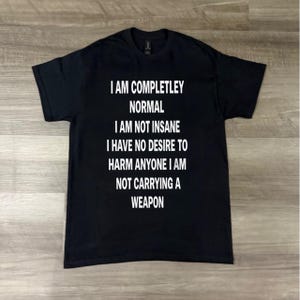 Puede incluir: Camiseta negra con texto blanco. El texto dice: "SOY COMPLETAMENTE NORMAL NO ESTOY LOCO NO TENGO DESEO DE DAÑAR A NADIE NO LLEVO ARMAS."