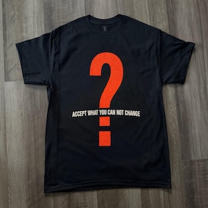 Puede incluir: Camiseta negra con un gran signo de interrogación naranja. El texto blanco debajo dice "ACCEPT WHAT YOU CAN NOT CHANGE". La camiseta se muestra sobre una superficie de madera.
