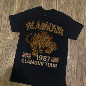 Puede incluir: Camiseta negra con un gráfico de leopardo y la palabra "GLAMOUR" en una fuente con estampado de leopardo. Debajo del leopardo está el texto "1987 GLAMOUR TOUR" con las banderas británica y estadounidense.