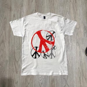 Puede incluir: Una camiseta blanca de manga corta con un gráfico grande de un símbolo de la paz rojo desgastado centrado en el pecho, rodeado por varios símbolos de la paz más pequeños en negro y gris con aspecto desgastado y de tamaños variados.