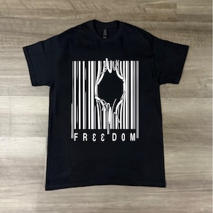 May include: Black t-shirt with a white graphic design. The design features a barcode-like pattern with a stylized image in the center and the word "FREEDOM" in white block letters at the bottom. The shirt is displayed on a wooden surface.