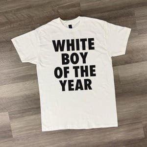 Op de afbeelding: Wit T-shirt met de tekst "WHITE BOY OF THE YEAR" in grote, zwarte hoofdletters. Het shirt is gemaakt van witte stof en heeft een ronde hals.