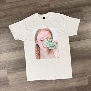Puede incluir: Camiseta de color crema con una ilustración en acuarela de una persona llorando mientras sostiene un fajo de dinero. La persona tiene el pelo largo y trenzado y se ven lágrimas. La obra de arte está centrada en la camiseta.