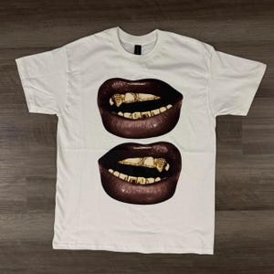 May include: White t-shirt featuring a graphic of two sets of lips with dark brown lipstick and gold teeth. The shirt is made of a soft, comfortable material.
