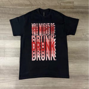 Puede incluir: Camiseta negra con el texto "YOU MIGHT BE DRUNK" en un diseño superpuesto rojo y blanco. El gráfico está alineado verticalmente y centrado en la camiseta. La camiseta es de material suave.