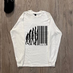 May include: A white long-sleeve t-shirt featuring a black graphic print on the chest. The design depicts the evolution of humans transitioning into a vertical barcode with the numbers 0 1234 78 printed underneath the barcode lines.