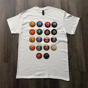 Puede incluir: Camiseta blanca con un diseño de bolas de billar numeradas del 1 al 15. Las bolas están dispuestas en una cuadrícula, cada una con un color y número diferente. La camiseta está hecha de un material suave.