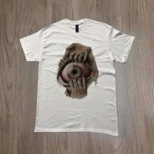 Może przedstawiać: Biała koszulka z surrealistyczną grafiką szczegółowego oka trzymanego otwartego przez dłonie z pierścieniami. Oko ma brązową tęczówkę i czerwono-różowe otoczenie. Koszulka jest wykonana z miękkiego materiału.