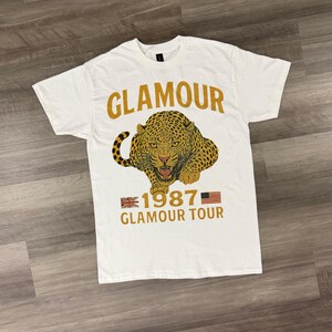Puede incluir: Camiseta blanca con un estampado de leopardo dorado y la palabra "GLAMOUR" encima. Debajo del leopardo está el texto "1987 GLAMOUR TOUR" con una bandera británica y estadounidense a cada lado.