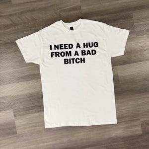 Può includere: T-shirt bianca con la scritta nera "I NEED A HUG FROM A BAD BITCH". La maglietta ha un girocollo ed è realizzata in un materiale morbido.