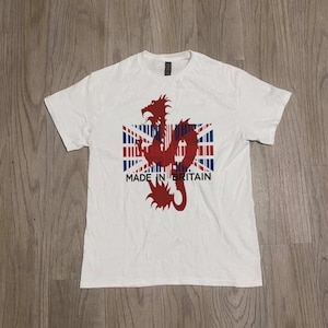 Puede incluir: Camiseta blanca con un diseño de dragón rojo sobre una bandera de la Union Jack. El texto "MADE IN BRITAIN" está impreso debajo. La camiseta está hecha de un material suave.