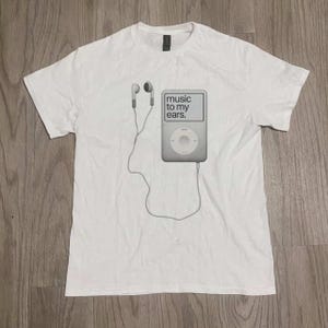 Könnte beinhalten: Weißes T-Shirt mit einem Graustufen-Grafikdesign eines MP3-Players und Kopfhörern, die durch ein Kabel verbunden sind. Der Text "music to my ears" ist auf dem Bildschirm des Players zu sehen. Schlichtes, lässiges Design.