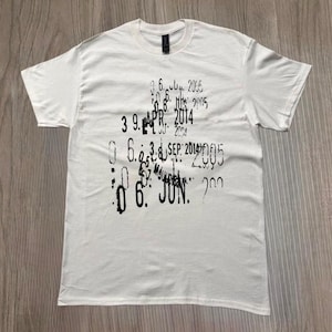 Puede incluir: Camiseta blanca con cuello redondo y mangas cortas. La camiseta presenta una impresión negra desgastada de fechas y números. Las fechas incluyen "ABR 2014" y "JUN". La camiseta está hecha de un material suave.