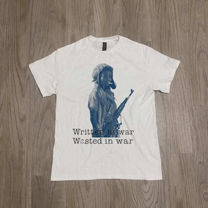 Puede incluir: Camiseta blanca con un gráfico azul de una persona con un rifle, con un tocado y fumando un cigarrillo. El texto "Written in war Wasted in war" está impreso debajo.