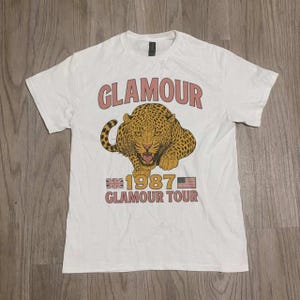 Puede incluir: Camiseta blanca con un diseño gráfico que presenta la palabra "GLAMOUR" sobre una ilustración de leopardo. Debajo, las palabras "1987 GLAMOUR TOUR" con banderas británica y estadounidense.