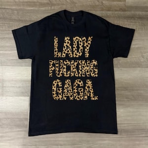 Puede incluir: Camiseta negra con el texto "LADY FUCKING GAGA" en un diseño de estampado de leopardo. La camiseta es de cuello redondo y parece estar hecha de un material suave.