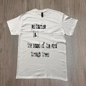Puede incluir: Una camiseta blanca con la palabra "psithurism" y su definición impresa en negro. La definición dice "el sonido del viento a través de los árboles."