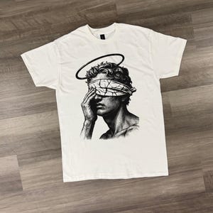 Puede incluir: Camiseta blanca con una ilustración en blanco y negro de un busto con un halo y una venda en los ojos. El gráfico es un dibujo detallado de una persona cubriéndose los ojos. La camiseta está tendida sobre una superficie de madera.