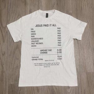 Puede incluir: Camiseta blanca con la frase "JESUS PAID IT ALL" y una lista de pecados marcados como "PAGADO". La camiseta incluye el texto "CANTIDAD A PAGAR 0,00" y un versículo bíblico. Prenda casual de temática religiosa.