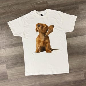 以下が含まれることがあります： 白いTシャツ。茶色の犬がイヤホンをして目を閉じています。犬は耳を立てて座っており、イヤホンのコードが見えます。このシャツは柔らかい素材で作られています。