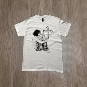 Puede incluir: Camiseta blanca con una ilustración en blanco y negro de una persona sentada y una cruz. La camiseta gráfica tiene cuello redondo y mangas cortas. La ilustración representa a una persona sentada con las rodillas en el pecho.