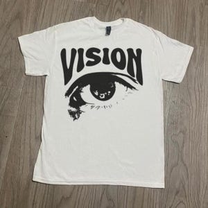 Peut inclure: T-shirt blanc avec un graphisme noir représentant un œil et le mot "VISION" au-dessus. Le graphisme de l'œil est détaillé avec des cils et une pupille. Le t-shirt est en matière souple.