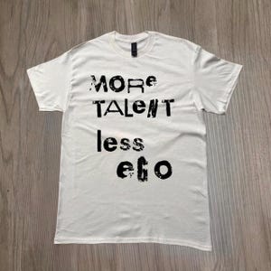 Puede incluir: Camiseta blanca con el texto negro "MORE TALENT less ego". La camiseta tiene mangas cortas y cuello redondo. El texto está en un estilo grunge.