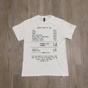 Puede incluir: Camiseta blanca con la frase "JESUS PAID IT ALL" en negro, con un diseño que simula un recibo. El diseño enumera los pecados marcados como "PAGADO" e incluye un versículo bíblico. Diseño sencillo y basado en la fe.