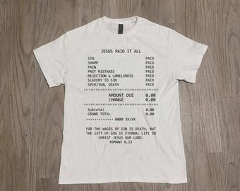 Könnte beinhalten: Ein weißes kurzärmeliges T-Shirt mit schwarzem Text im Kassenbon-Stil. Der Text lautet: JESUS PAID IT ALL, gefolgt von einer Liste als PAID markierter Punkte, darunter SIN, SHAME, PAIN, und endet mit dem Bibeltext ROMANS 6.23.