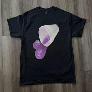 May include: Black t-shirt featuring a graphic of a white cup spilling purple liquid and ice cubes. The design is centered on the front of the shirt. The shirt is made of a soft material.