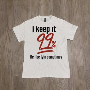 Könnte beinhalten: Weißes T-Shirt mit dem Text "I keep it 99%" in Schwarz und Rot. Der Satz "Bc i be lyin sometimes" ist unter der Zahl aufgedruckt. Das Shirt besteht aus einem weichen Material.