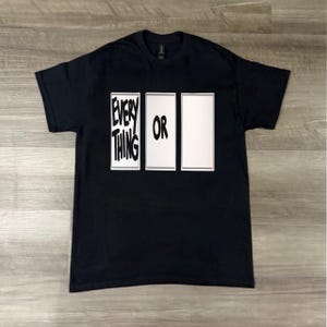 Puede incluir: Camiseta negra con las palabras "EVERYTHING OR" en blanco y negrita. El texto está dispuesto en tres rectángulos verticales. La camiseta se muestra sobre una superficie clara con vetas de madera.
