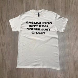 Puede incluir: Camiseta blanca con el texto negro "GASLIGHTING ISN'T REAL YOU'RE JUST CRAZY". La camiseta tiene mangas cortas y cuello redondo. La camiseta se muestra sobre una superficie de vetas de madera clara.