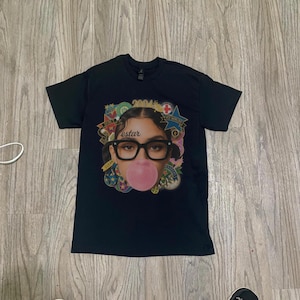 Puede incluir: Camiseta negra con un gráfico de una persona con gafas soplando una burbuja de chicle. La palabra "estar" está impresa en la camiseta. El gráfico está rodeado de varias insignias coloridas y el número "2004".