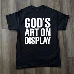 Puede incluir: Camiseta negra con el texto blanco "GOD'S ART ON DISPLAY". La camiseta es de un material suave y tiene cuello redondo. El texto está centrado en la parte delantera de la camiseta.