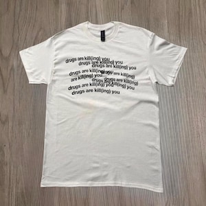 Puede incluir: Camiseta blanca con el texto "drugs are kill(ing) you" repetido en un patrón diagonal. La camiseta tiene mangas cortas y cuello redondo.