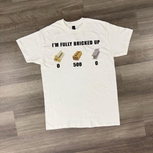 Puede incluir: Camiseta blanca con el texto "I'M FULLY BRICKED UP" e ilustraciones de troncos, un ladrillo y una viga metálica. El gráfico del ladrillo tiene el número 500 debajo. Los otros gráficos tienen el número 0 debajo.