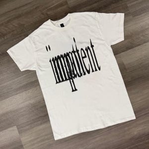 Puede incluir: Camiseta blanca con la palabra "impatient" en fuente negra estilizada. La camiseta tiene cuello redondo y mangas cortas. El texto está centrado en la parte delantera de la camiseta.