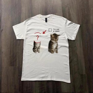May include: White t-shirt featuring a graphic of a cat with a question mark above its head and a pixelated cat. Text above the pixelated cat reads "I'm not a robot".