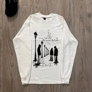 May include: A white long-sleeve shirt featuring a black ink-style graphic of figures standing along a path of street lamps. The design includes text that reads: no, my not tine, is me repeated, ay, the world that was, no, tine, ated, ay, world that was.