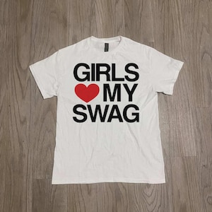 Puede incluir: Camiseta blanca con el texto "GIRLS ❤️ MY SWAG" en letras negras y un corazón rojo. La camiseta es de material suave y tiene mangas cortas.