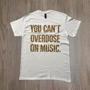 May include: White t-shirt with the text "YOU CAN'T OVERDOSE ON MUSIC" in a leopard print font. The shirt is made of a soft material and has a crew neck. The shirt is displayed on a wooden surface.