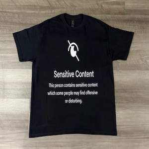 Op de afbeelding: Zwart T-shirt met de tekst "Sensitive Content" en een afbeelding erboven. De tekst hieronder luidt: "Deze persoon bevat gevoelige inhoud die sommige mensen aanstootgevend of storend kunnen vinden."