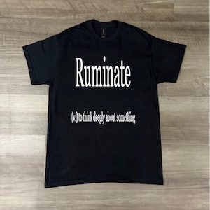 Könnte beinhalten: Schwarzes T-Shirt mit dem weißen Wort "Ruminate" und der Definition "(v.) to think deeply about something." Das Shirt liegt flach auf einer Holzoberfläche.