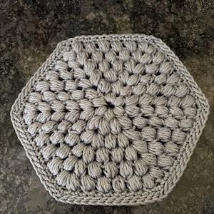 Puede incluir: Un salvamanteles hexagonal de ganchillo gris con un patrón texturizado en relieve. Los bordes están cuidadosamente acabados con un borde trenzado apretado. Este artículo hecho a mano es adecuado para la cocina.